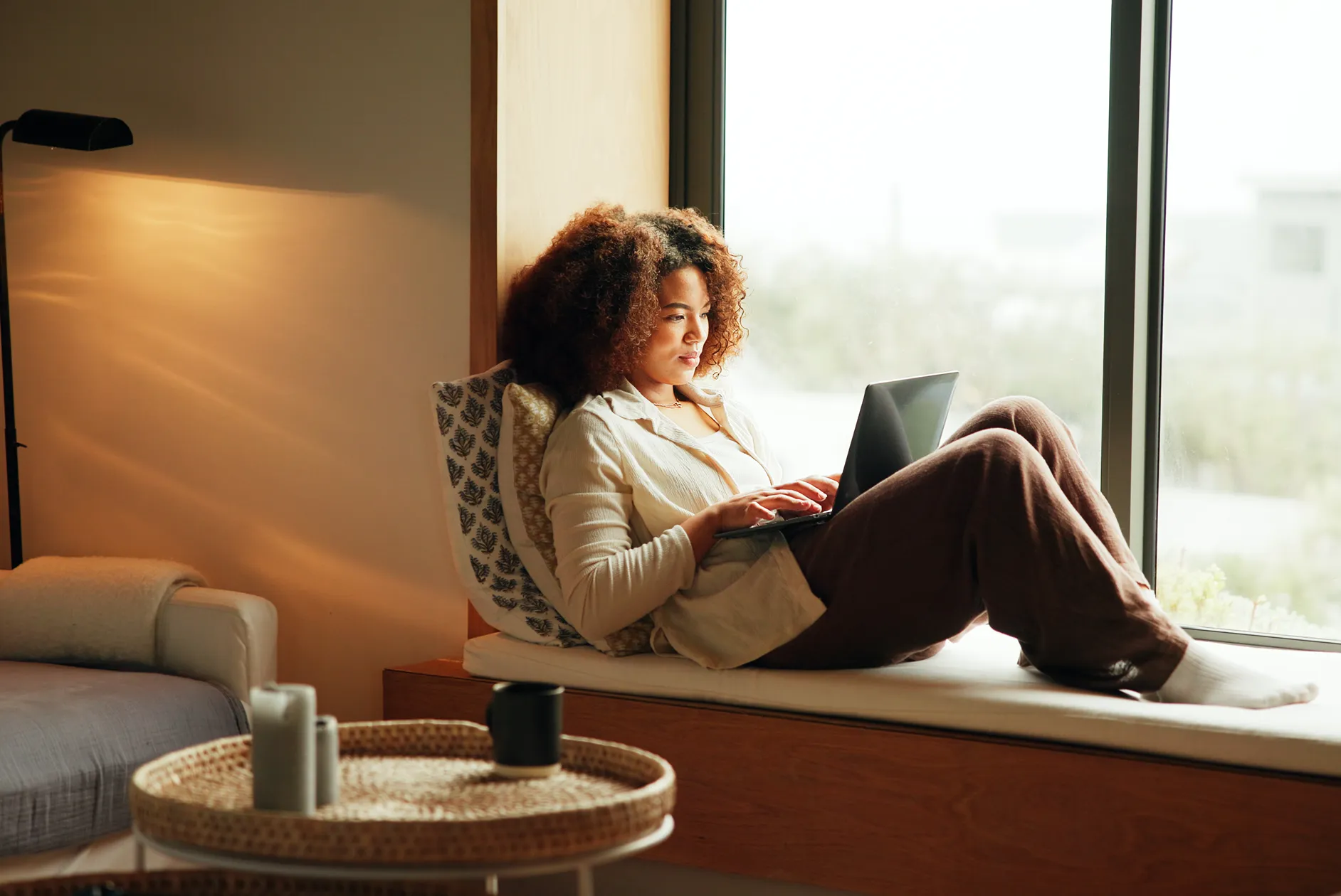 Woman with curly hair sitting on a window seat working on a laptop in a cozy, softly lit room.