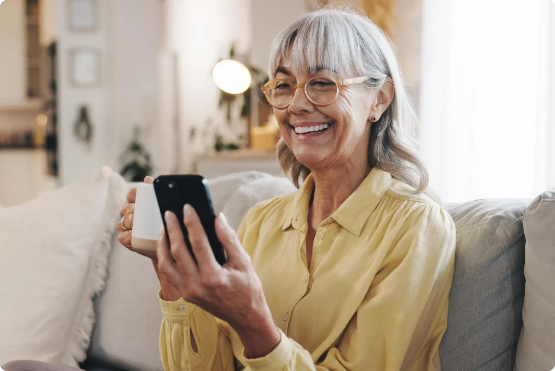 Smiling elderly woman with gray hair and glasses holding a smartphone in one hand and a white mug in the other while sitting on a couch.