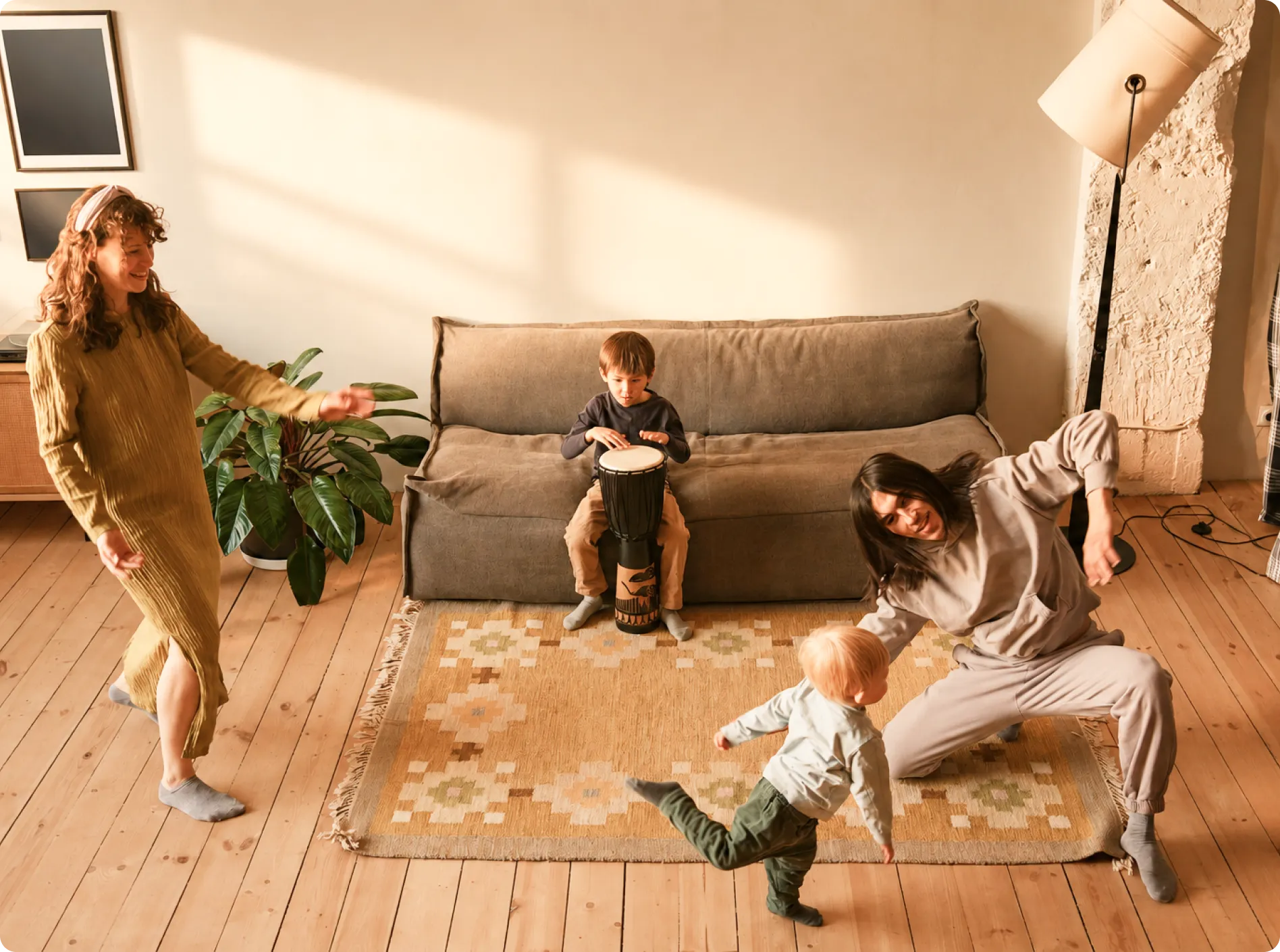 A child sitting on a sofa plays a drum while two adults and a toddler dance on a patterned rug.