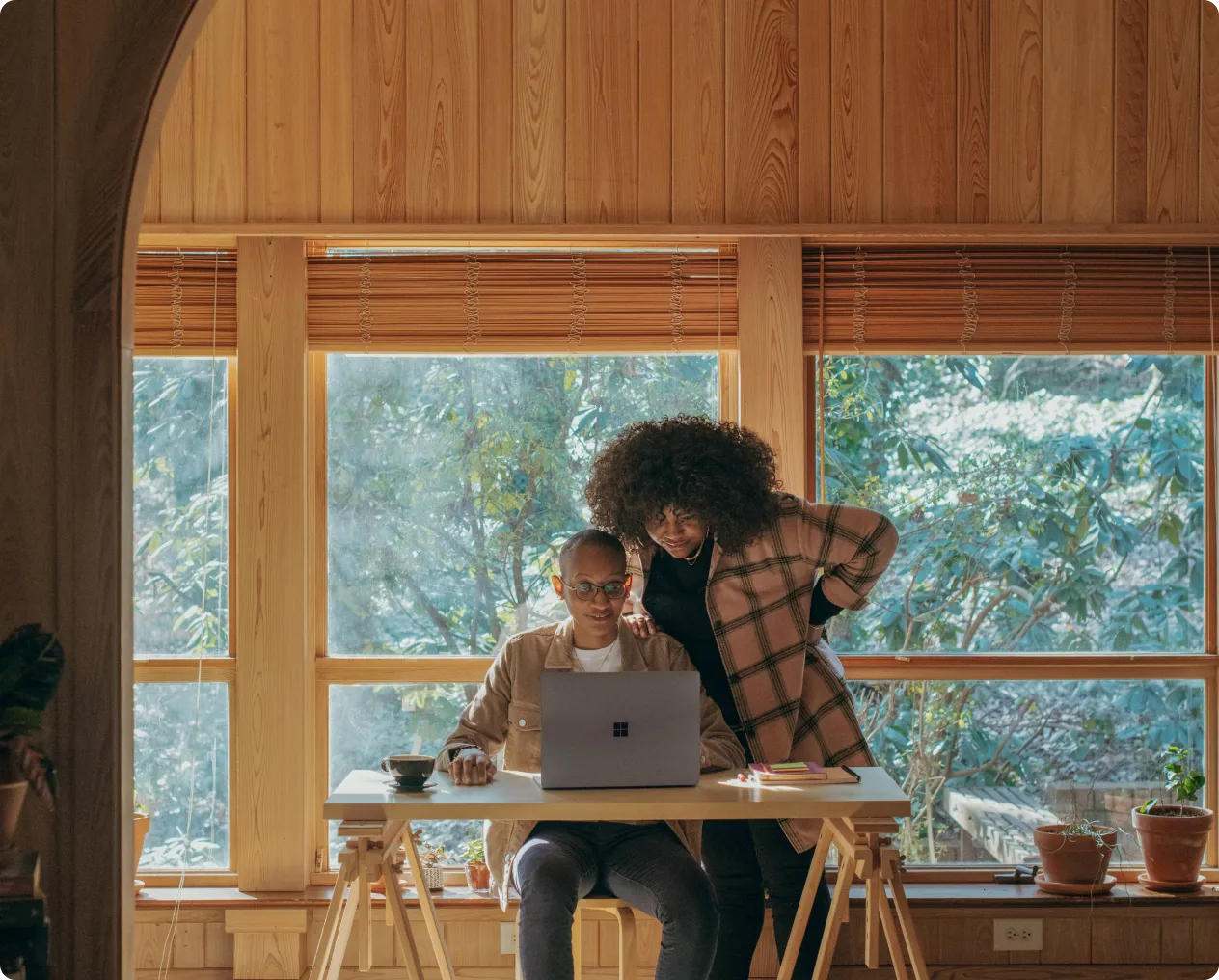 Two people working together at a desk with a laptop near large wooden-framed windows.
