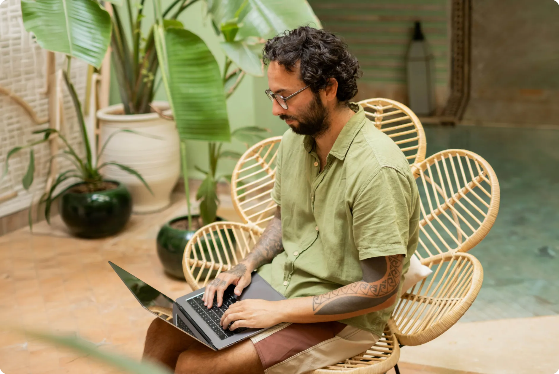 Man with tattoos wearing glasses and a green shirt using a laptop in a room with plants.
