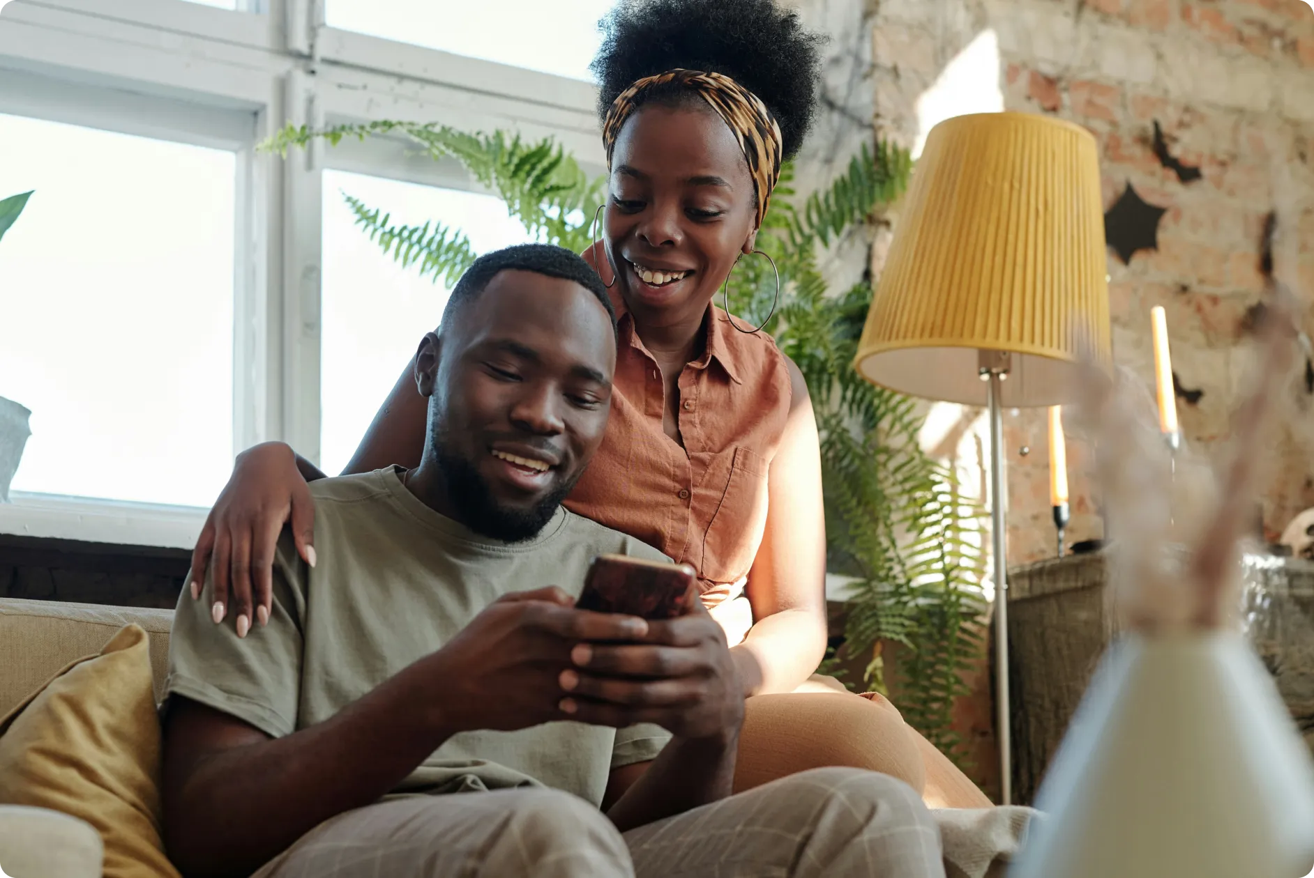 Smiling couple sitting on couch looking at phone with plants and lamp in background.