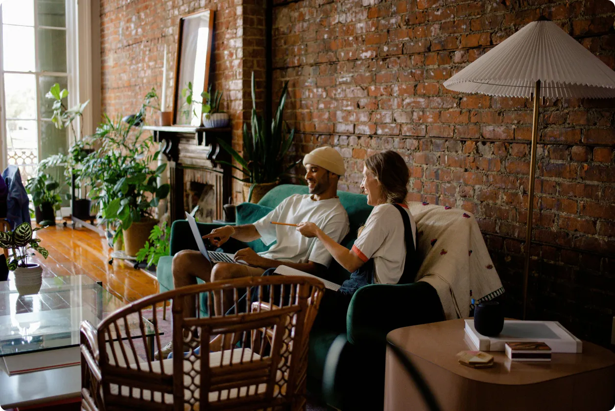 Two people sitting on a green couch with a laptop and notebook in a cozy, plant-filled living room.
