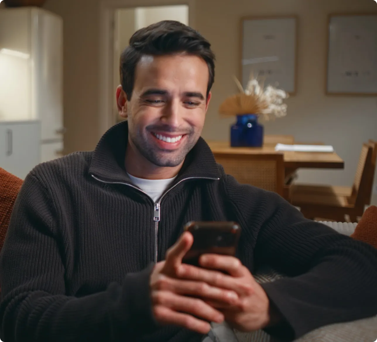Smiling man in black sweater looking at his smartphone while sitting on a couch at home.