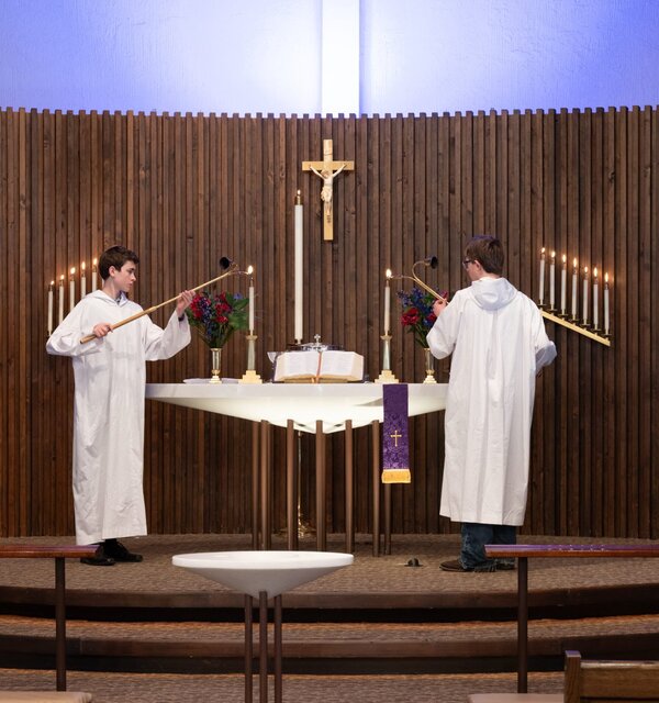 Two acolytes in white robes each light a candle on an altar. There is an open altar book between them, and a crucifix hangs behind them. A modern baptismal font sits in front of the chancel.
