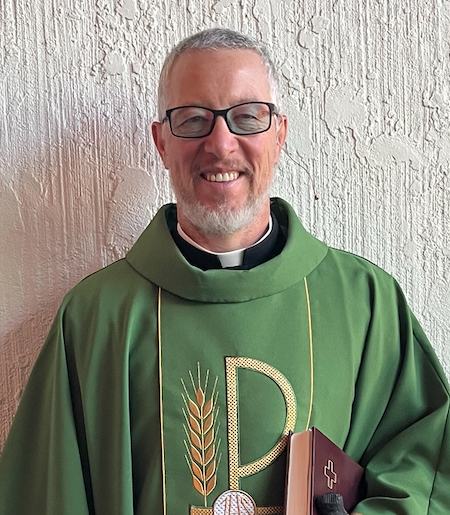 A headshot of Pastor Bill Beck of Rock of Ages Lutheran Church in Colorado Springs, CO. He's wearing a green chasuble with a large gold Chi Rho next to a grain of wheat, and is holding a maroon hymnal. He has black glasses and a white beard. 