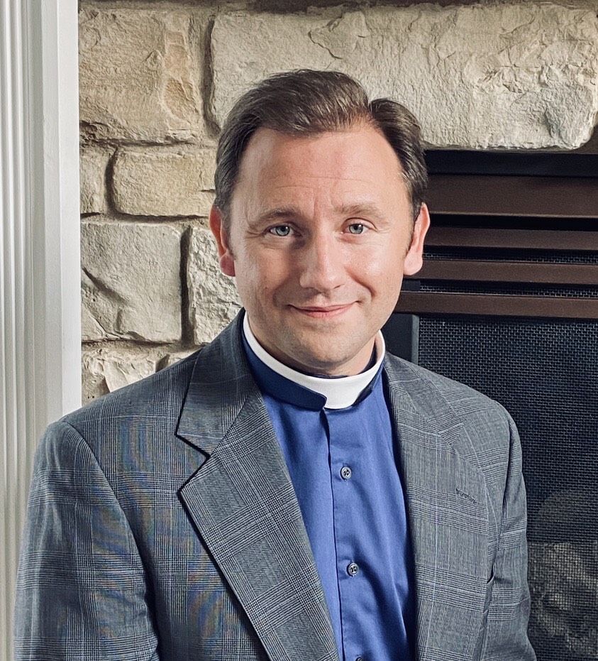Headshot of Pastor James Maxwell of Rock of Ages Lutheran Church in Colorado Springs, Colorado. He is wearing a grey suit jacket with a royal blue clerical shirt. He has a soft smile, brown hair, and is situated in front of a stone fireplace. 