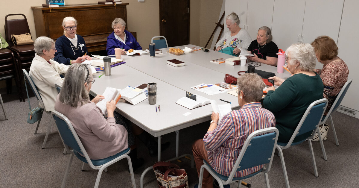 The ladies of Rock of Ages Lutheran Church in Colorado Springs, CO meet for their Thursday bible study. Many women are sitting around a table with bibles open and refreshments in front of them. 