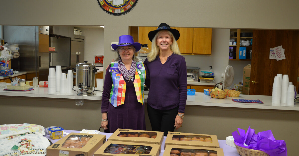 Two women in LWML in silly hats stand behind a table of treats at Rock of Ages Lutheran Church in Colorado Springs, CO 