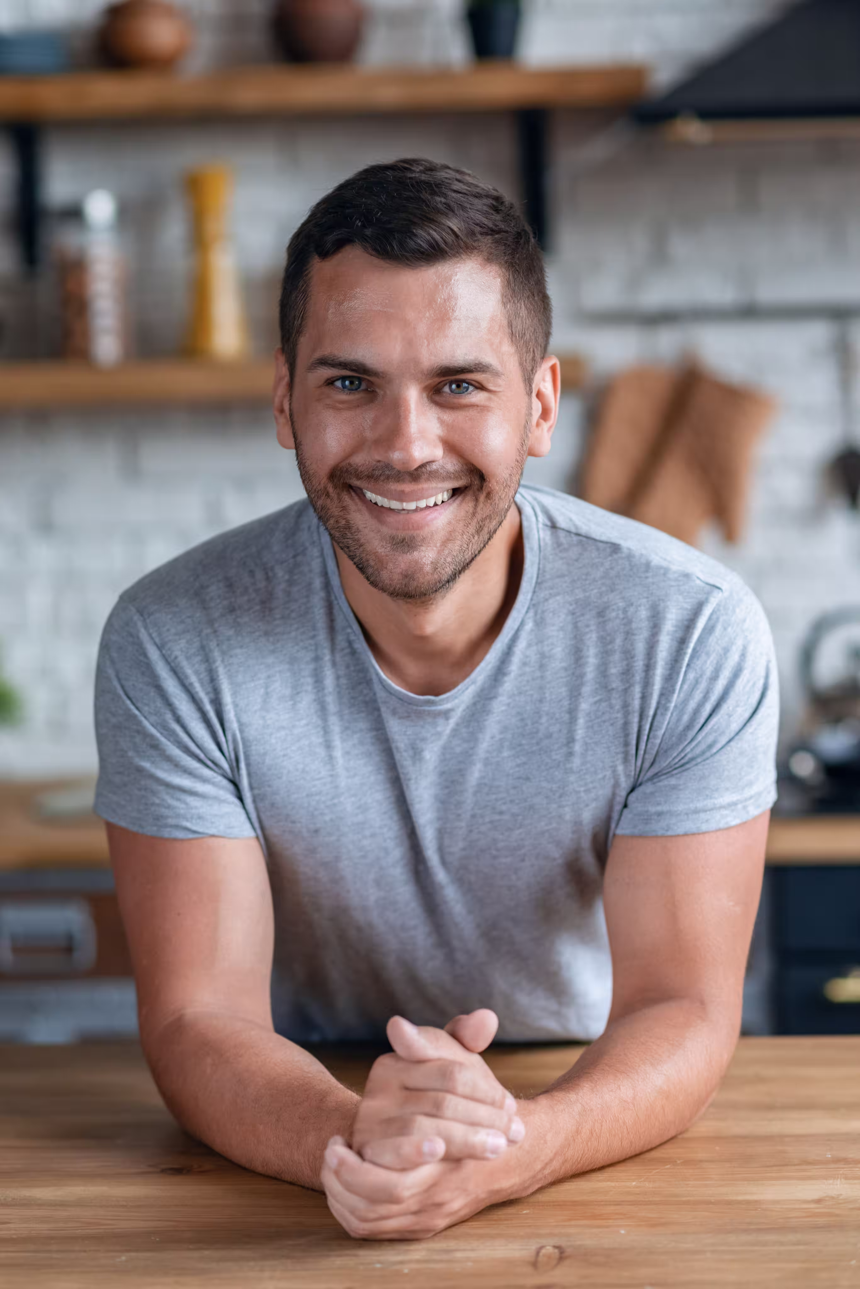 Smiling man in a gray t-shirt leaning on a wooden table, kitchen background with shelves and utensils.
