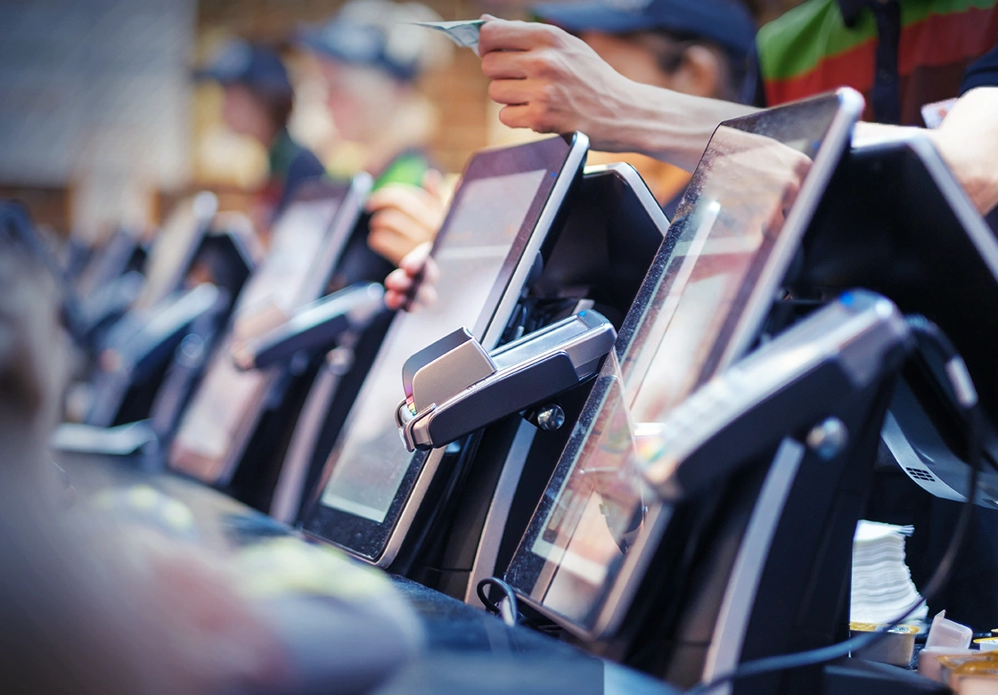 An image of a row of point of sale payment terminals and tablet POS devices, with a hand holding a credit card reaching over to pay.