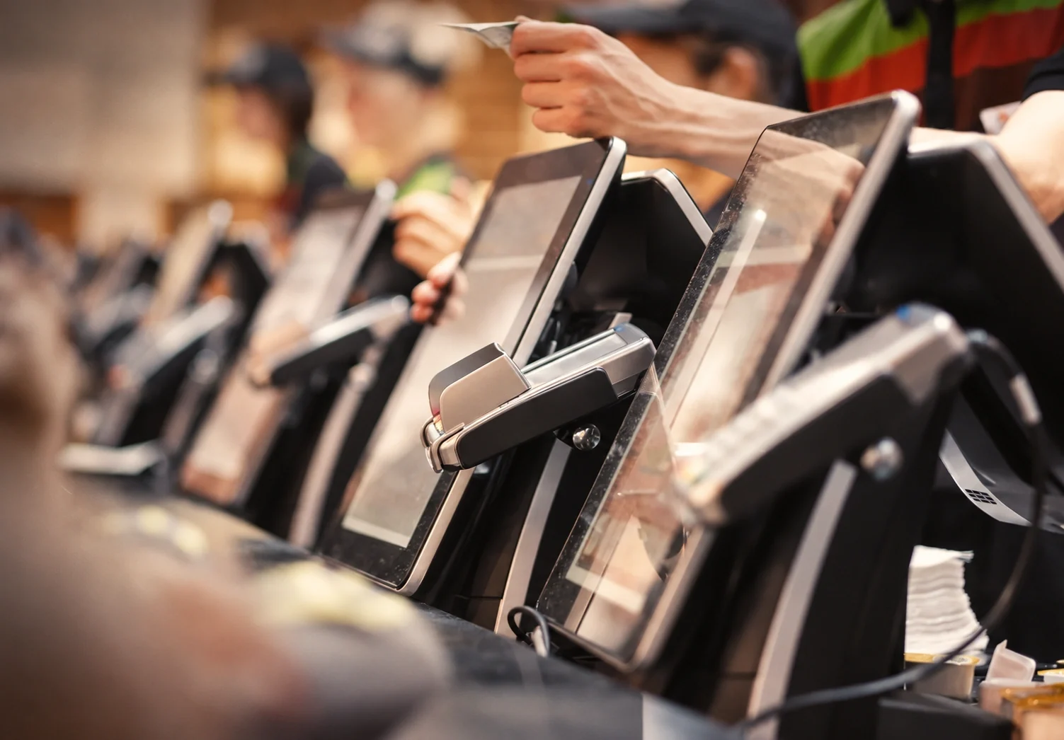An image of a row of point of sale payment terminals and tablet POS devices, with a hand holding a credit card reaching over to pay.