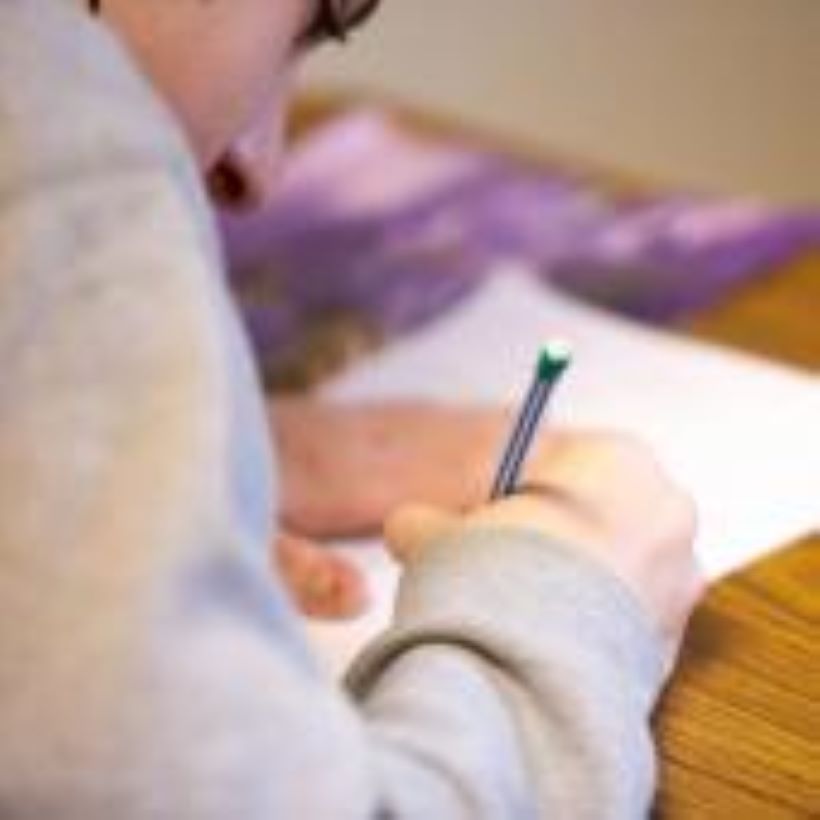 Niño con gafas escribiendo con un lápiz en un papel sobre una mesa de madera.