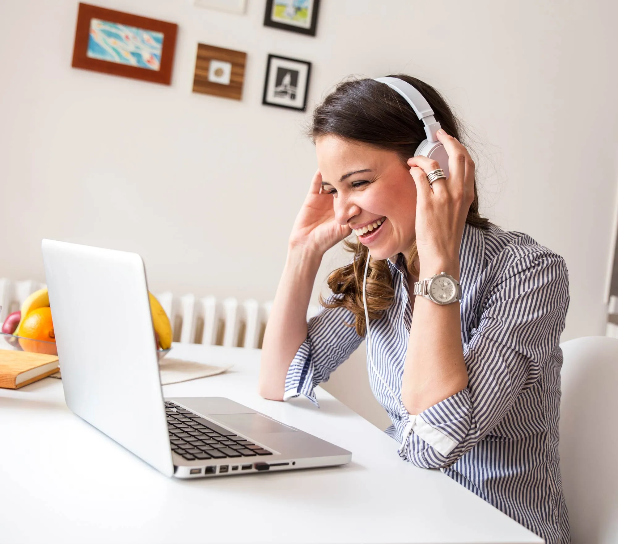 Mujer sonriente con auriculares blancos y camiseta a rayas mirando un portátil sobre una mesa blanca.