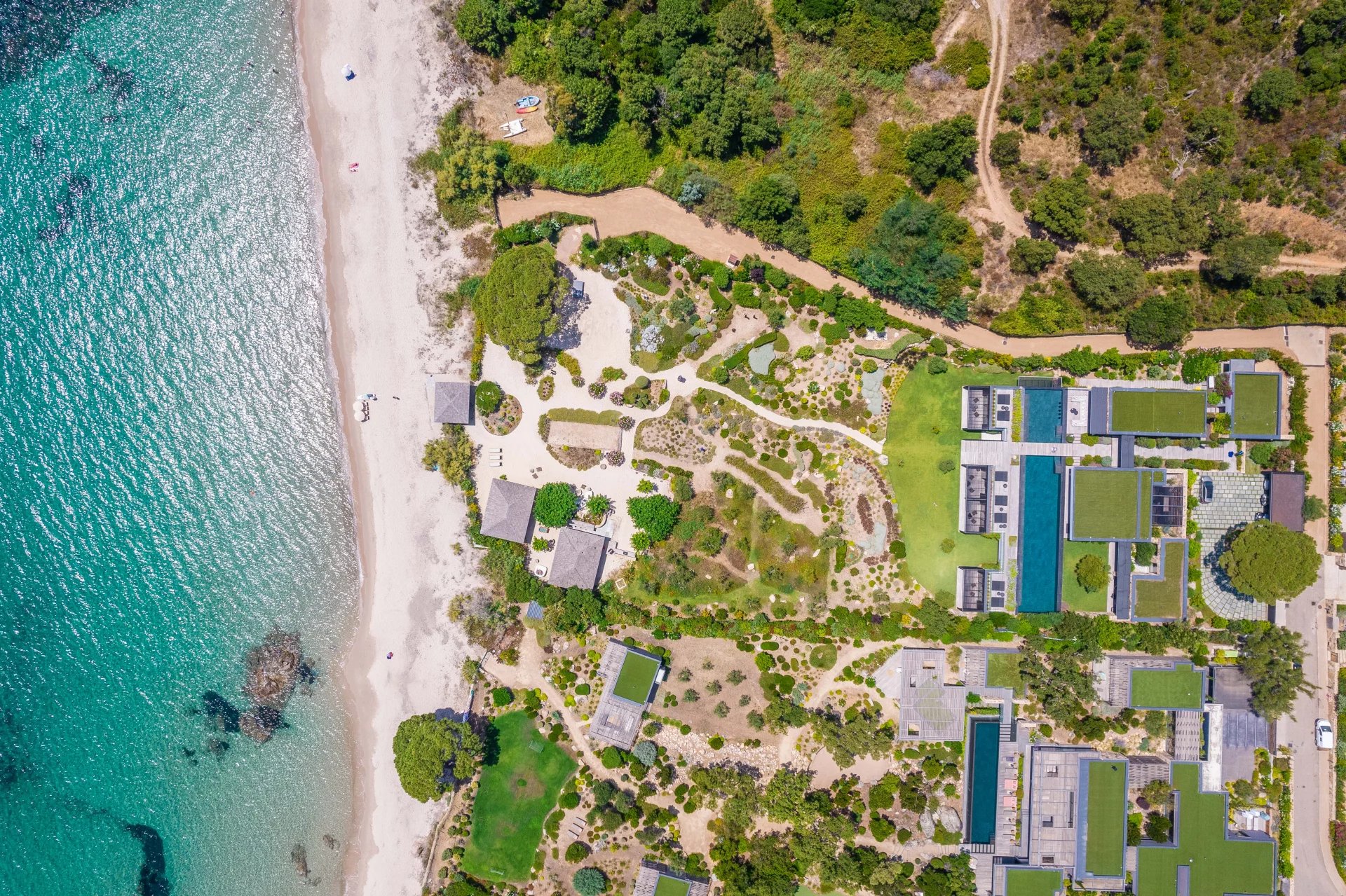 Vue aérienne d'une plage bordée d'une mer turquoise, avec des sentiers, des jardins et plusieurs bâtiments modernes dont certains ont des toits végétalisés.
