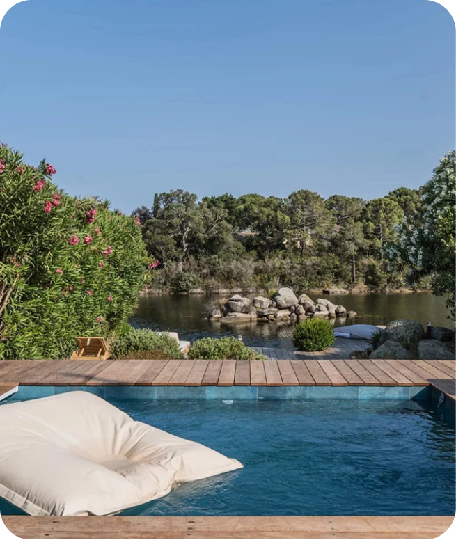 Piscine moderne avec un coussin flottant, entourée de terrasse en bois et vue sur un étang rocheux et des arbres verts sous un ciel bleu.