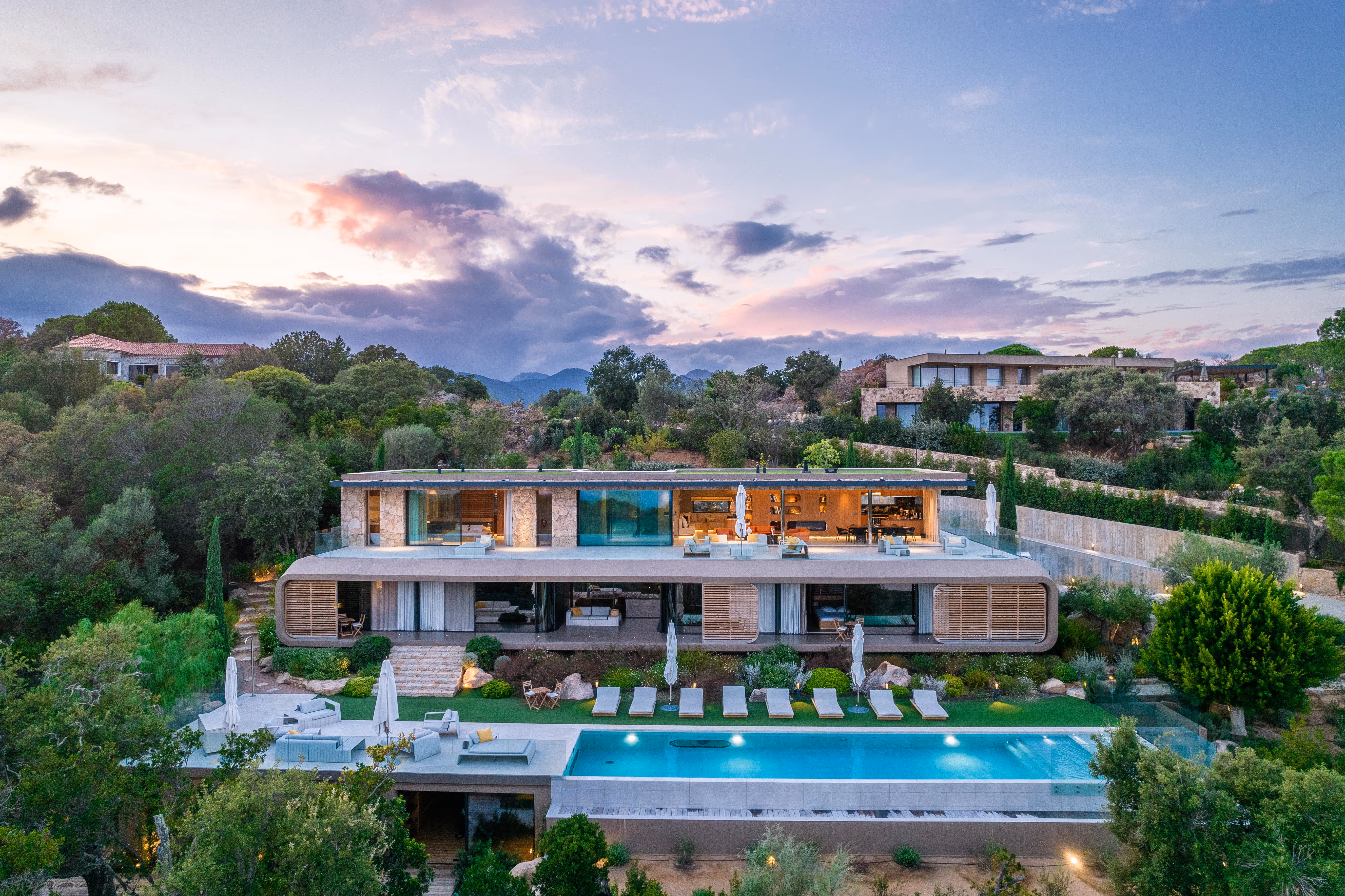 Vue panoramique d'une grande villa moderne avec terrasse, piscine et chaises longues au coucher du soleil entourée de végétation.