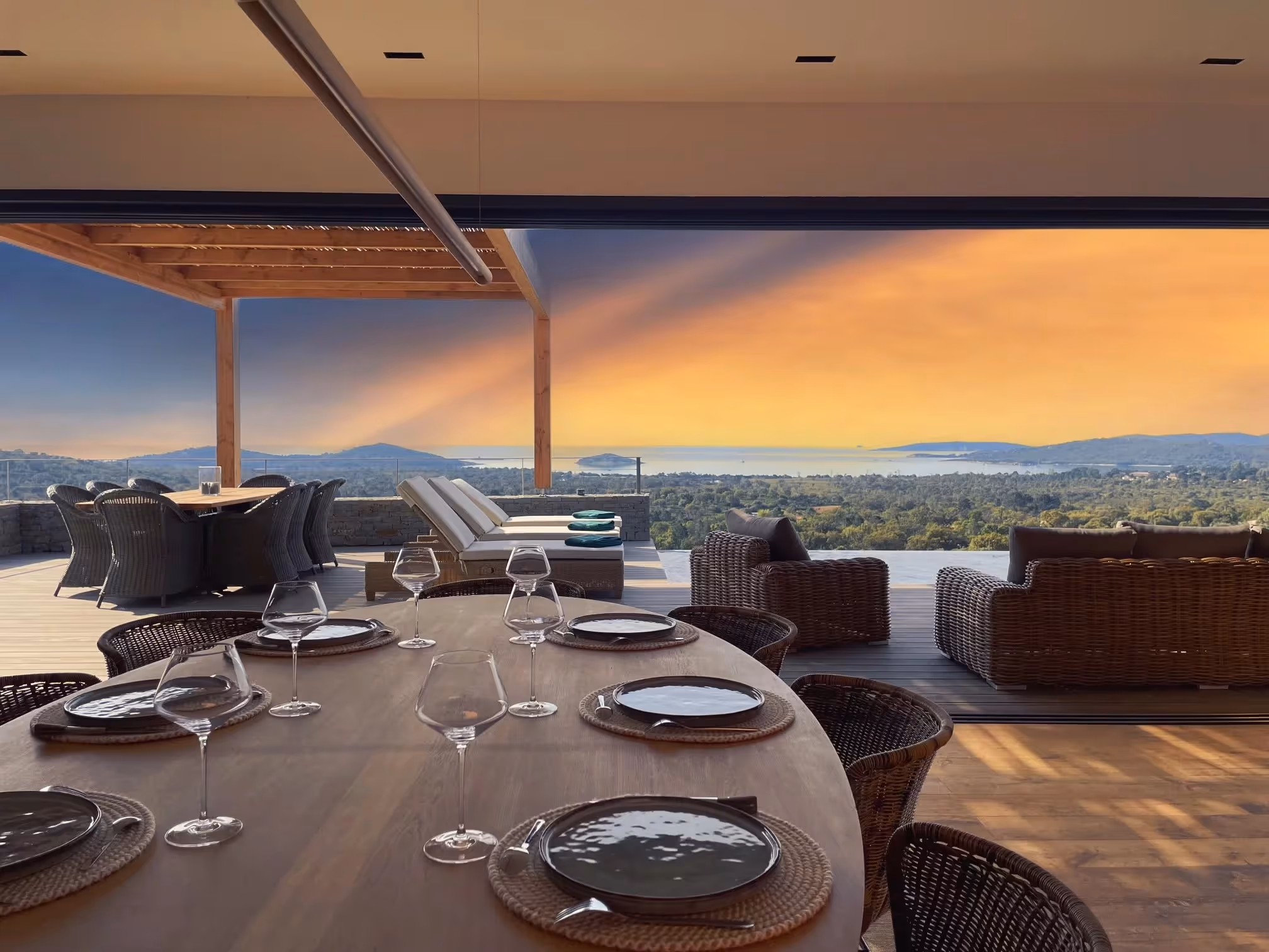 Vue d'une terrasse avec une grande table en bois dressée pour six personnes, des fauteuils en osier, des chaises longues blanches et un canapé extérieur avec vue sur une forêt et la mer au coucher du soleil.
