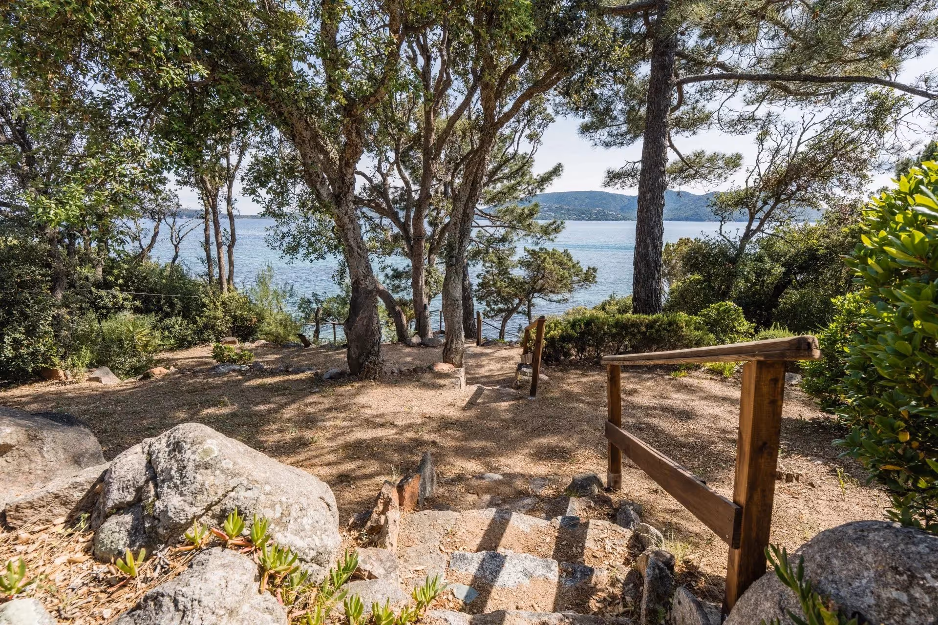 Escalier en pierre descendant vers une zone boisée avec vue sur un plan d'eau et des collines à l'arrière-plan.