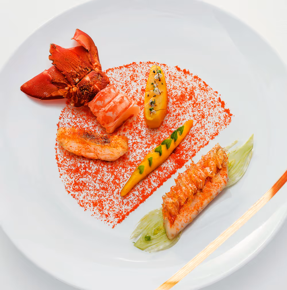 Assiette gastronomique avec morceaux de homard, purée décorée d'herbes, poudre rouge et une touche de sauce verte.