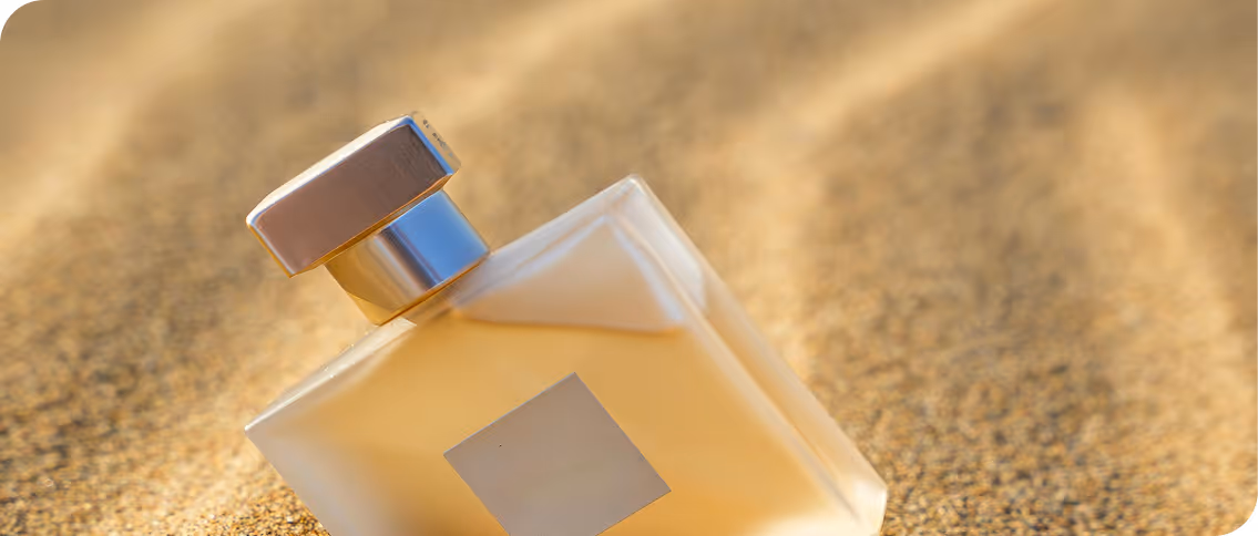Flacon de parfum carré avec un bouchon métallique repose sur du sable doré.