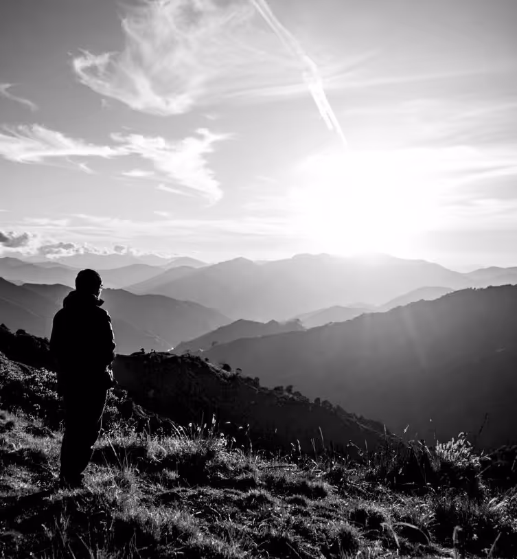 Silhouette of a person standing on a grassy hillside overlooking layered mountain ranges at sunrise.