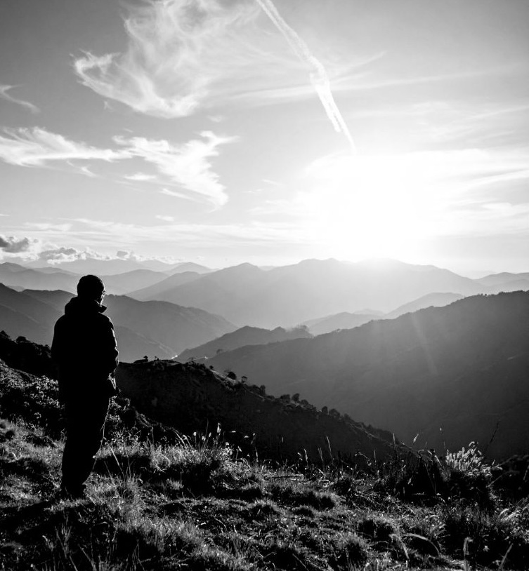 Silhouette of a person standing on a grassy hillside overlooking layered mountain ranges at sunrise.