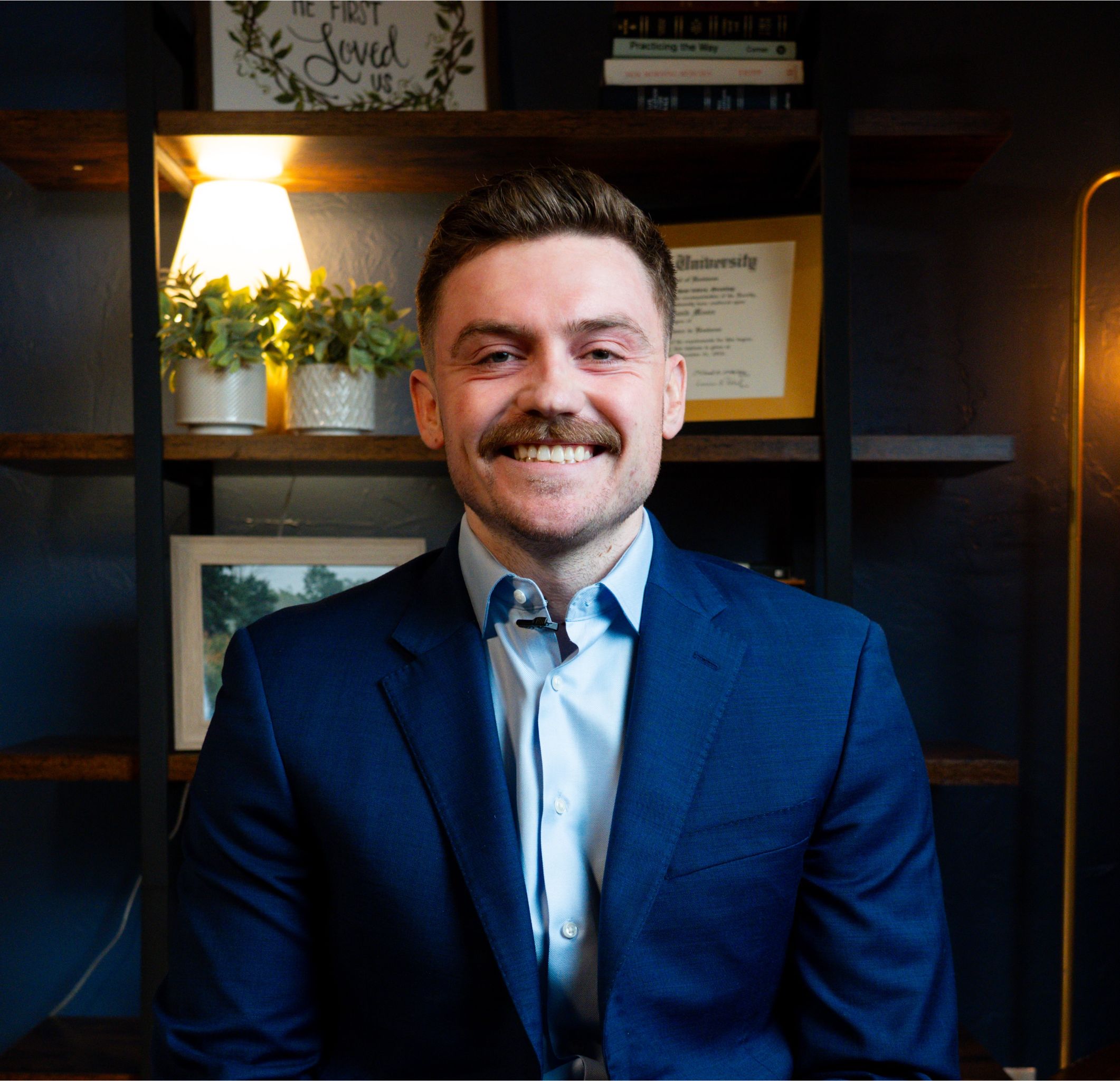 Smiling man with mustache wearing a blue suit jacket and light blue shirt sitting in front of a dark wooden shelf with plants, a lamp, framed photos, and books.