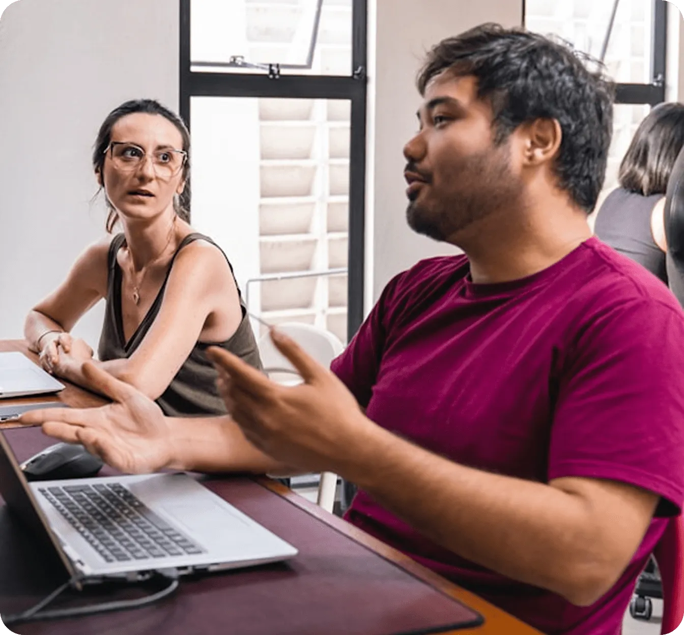 Un homme en t-shirt bordeaux parlant avec une femme portant des lunettes, assis devant des ordinateurs portables dans un bureau.