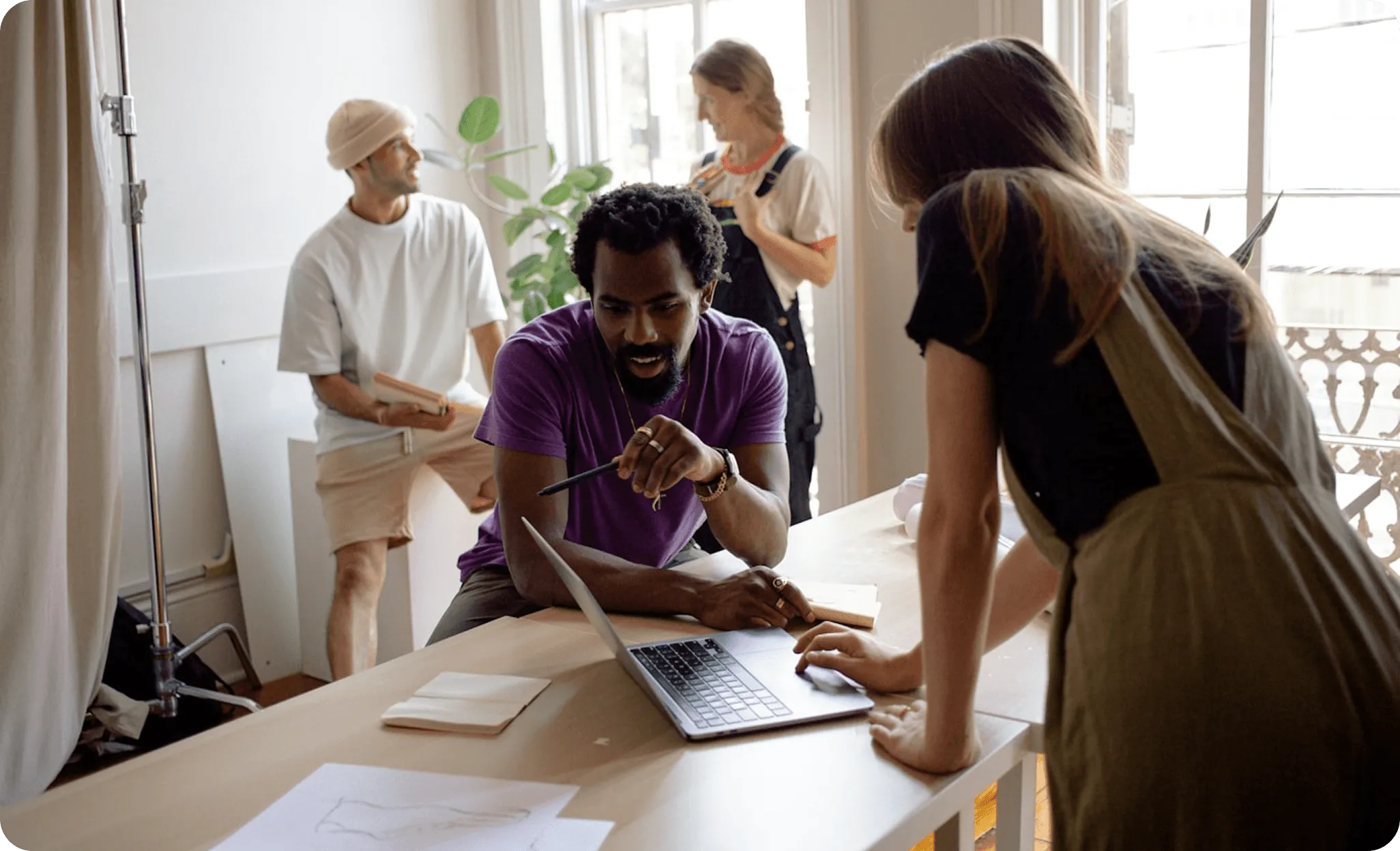 Quatre personnes collaborent dans un bureau lumineux, deux sont concentrées sur un ordinateur portable tandis que les deux autres discutent en arrière-plan.