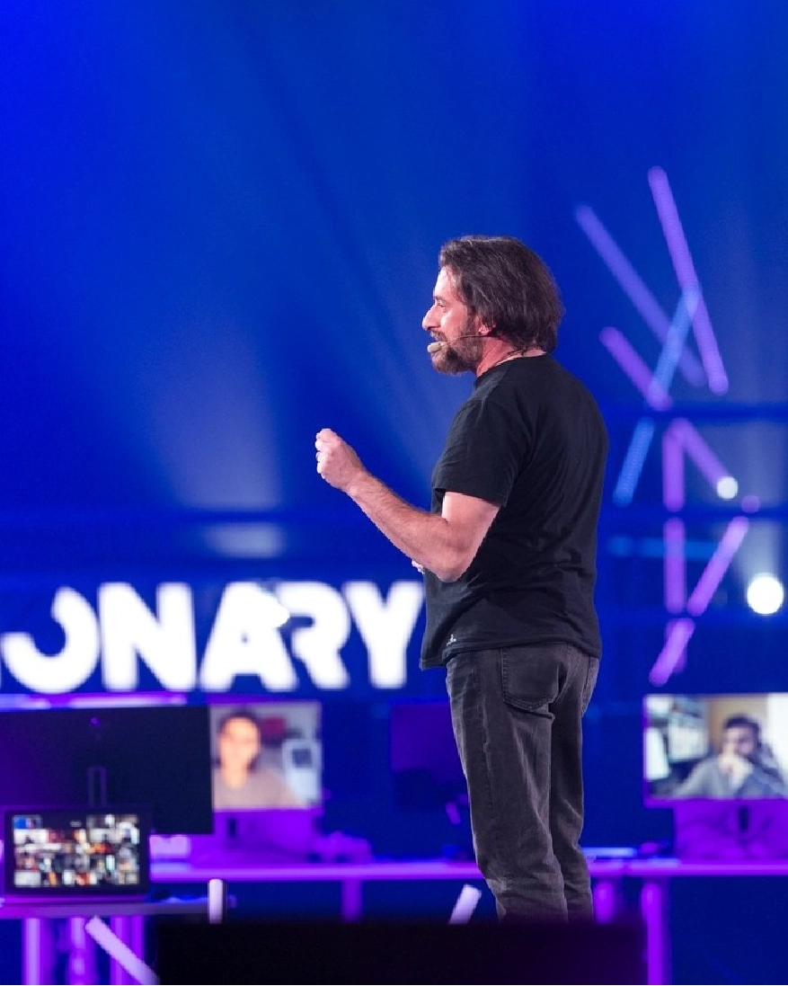 Uomo con microfono headset che parla su un palco con sfondo blu e schermi dietro di lui.