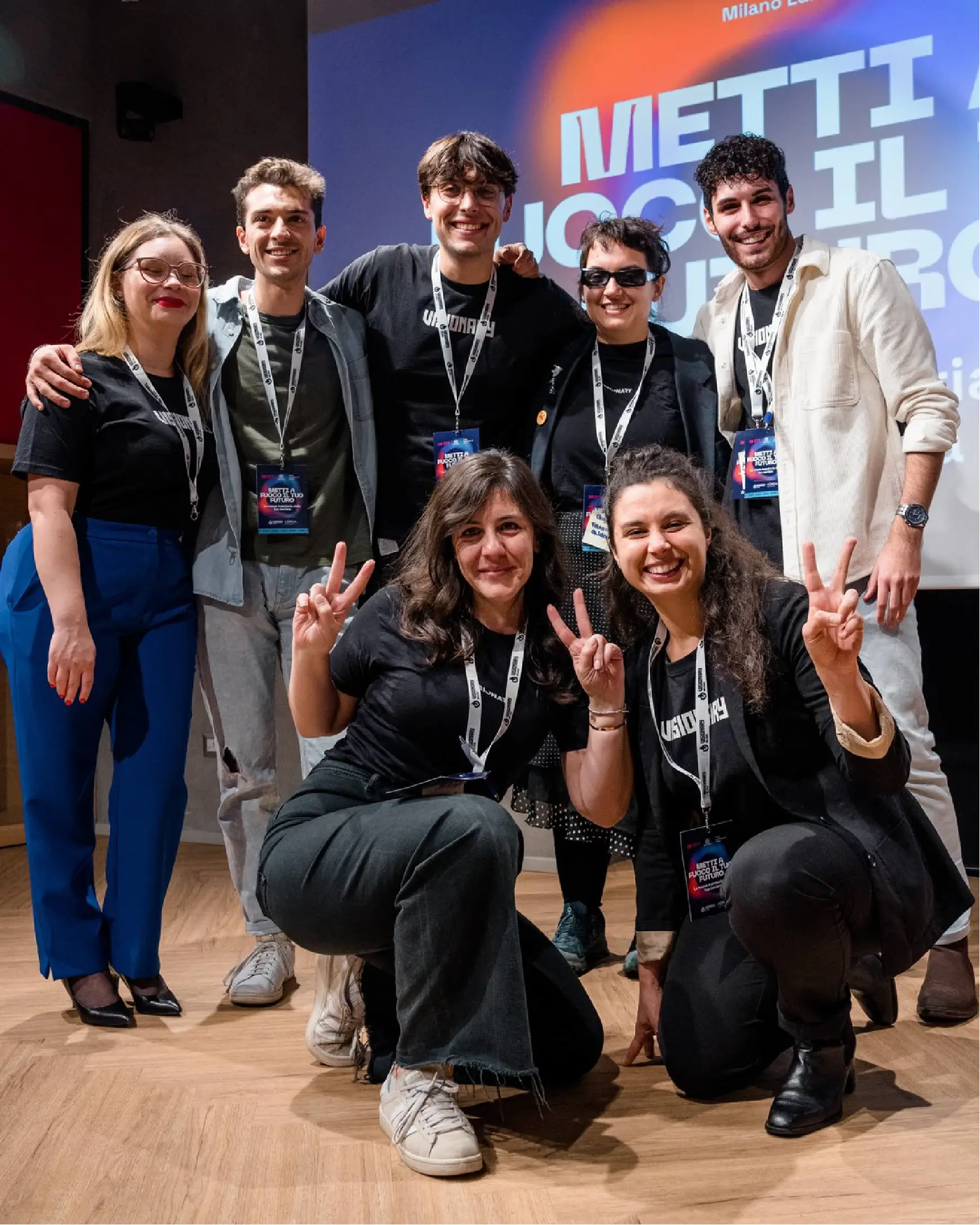 Gruppo di sette persone sorridenti con pass e magliette nere, due in ginocchio fanno il segno di pace con le dita.