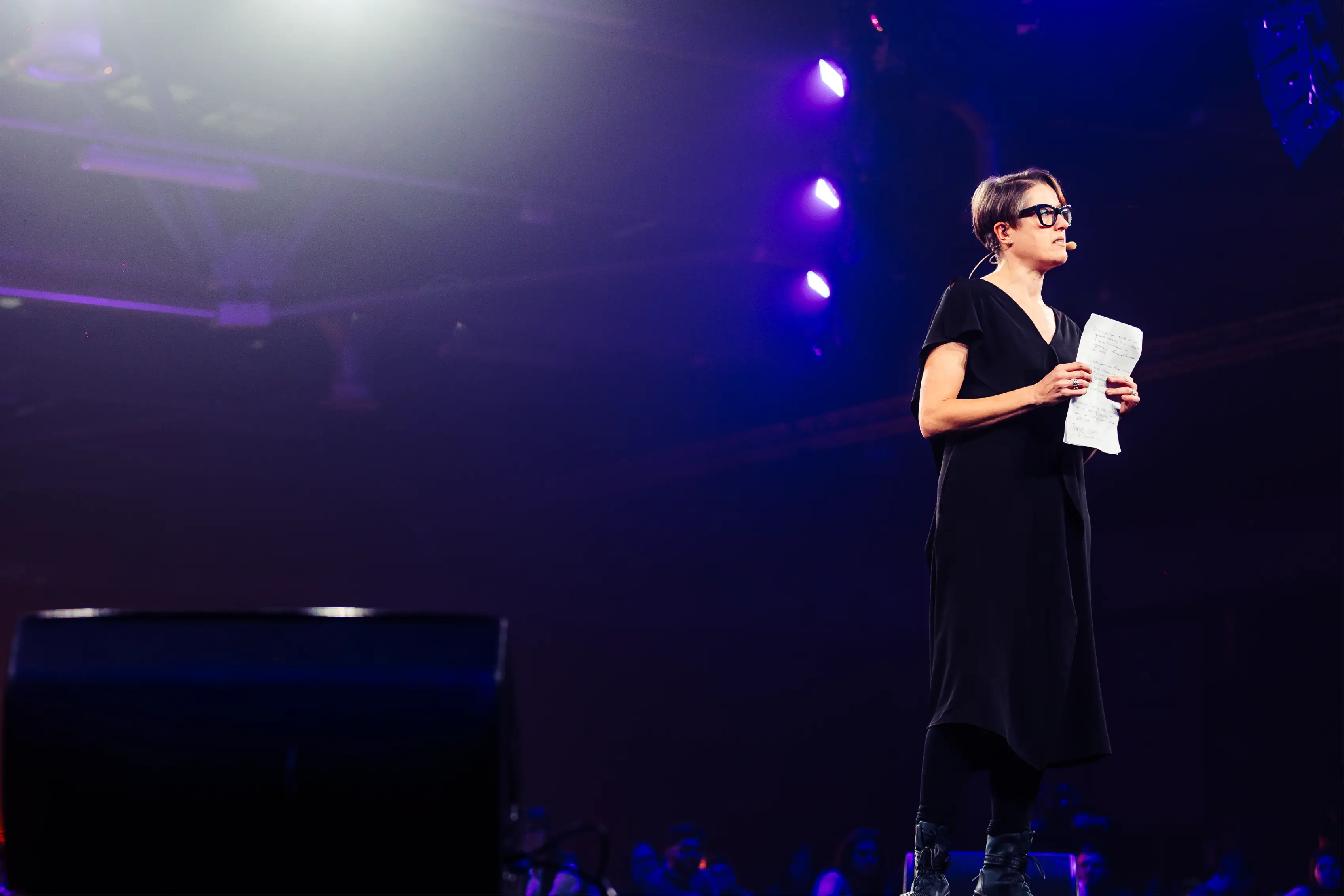 Donna con occhiali e microfono a collo che tiene foglio con appunti mentre parla su un palco illuminato.