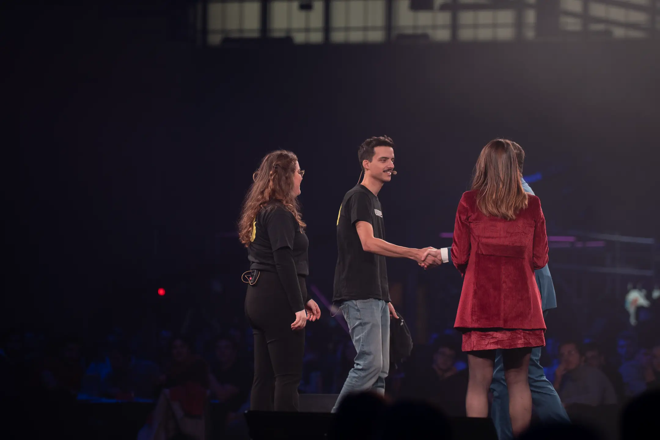 Quattro persone sul palco di un evento, con due che si stringono la mano, davanti a un pubblico in un ambiente scuro di un evento di Visionray Days.