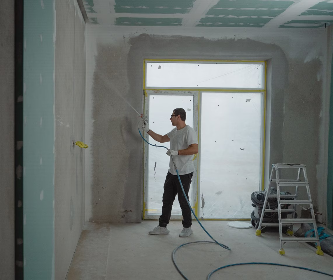 A worker wearing a white t-shirt and glasses uses a paint sprayer on the concrete walls of an unfinished interior room containing a large window and a stepladder.   