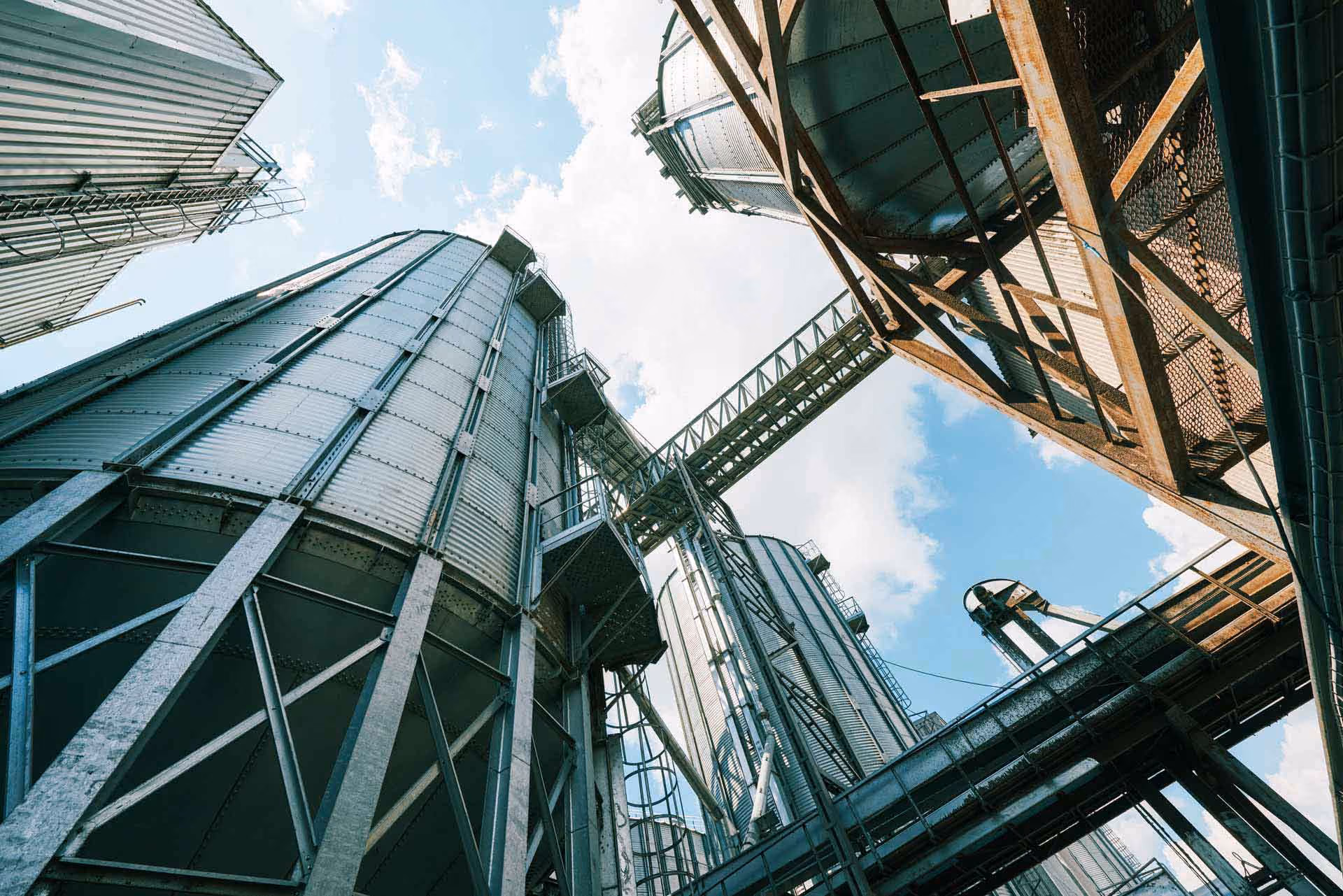 Vista para cima de vários silos metálicos altos sob céu azul com algumas nuvens.