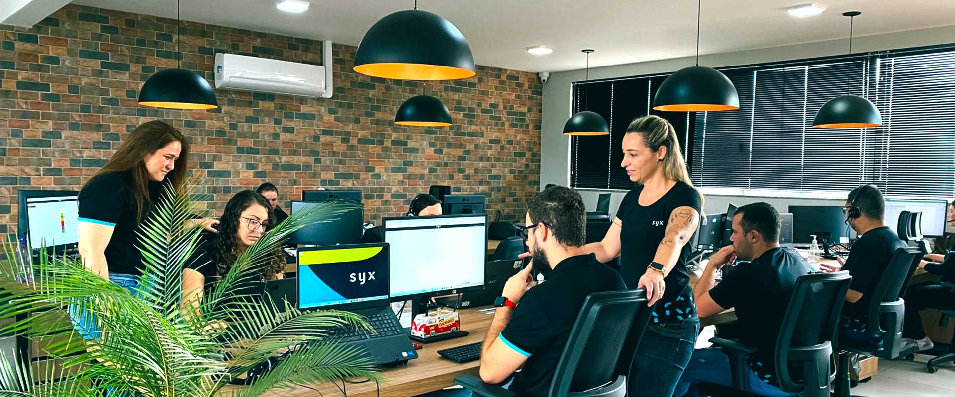 Equipe trabalhando em escritório moderno com computadores e plantas decorativas.