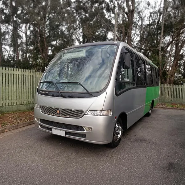 Ônibus prata com detalhe verde estacionado em rua com cercas e árvores ao fundo.