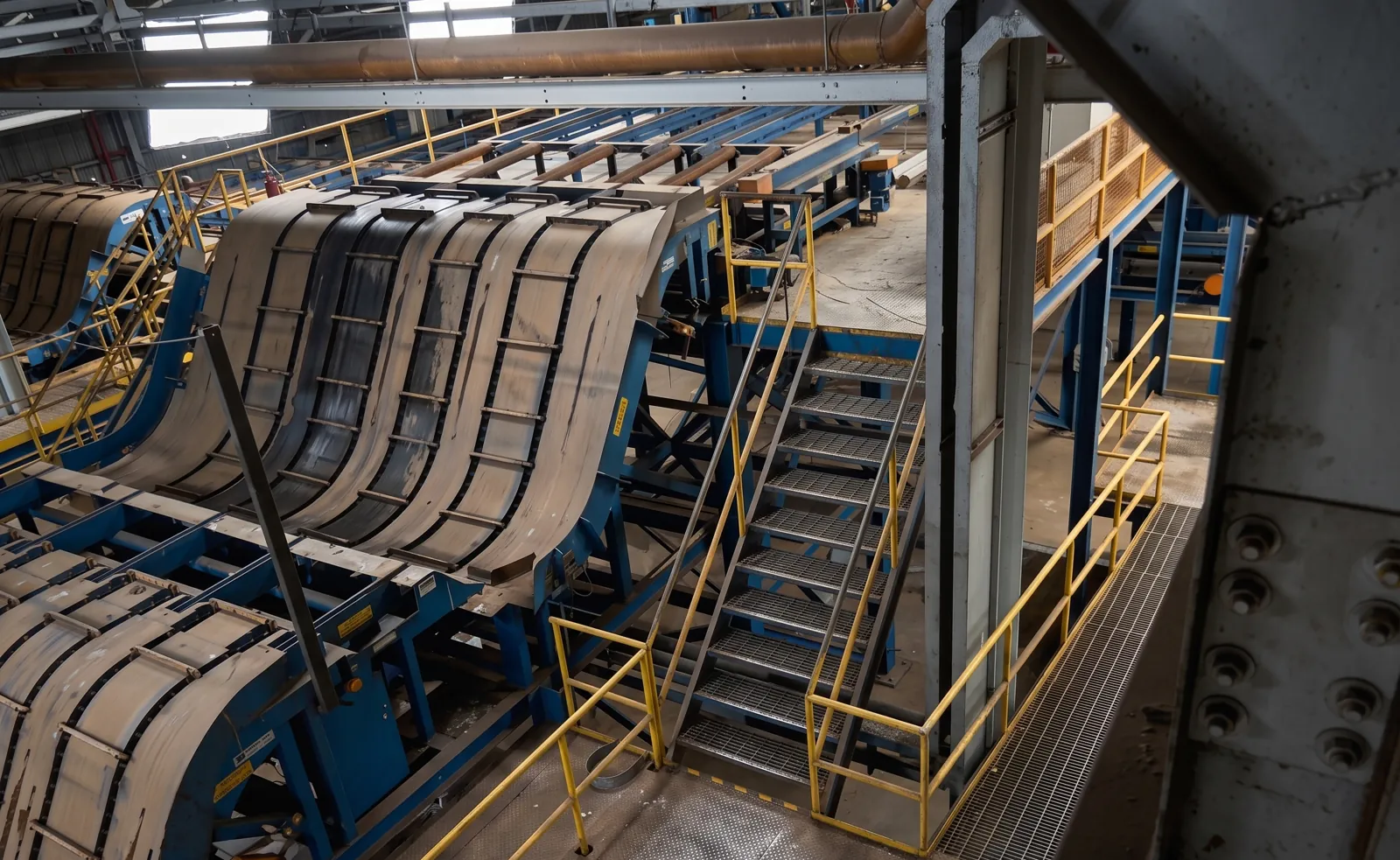 Esteiras transportadoras industriais azuis com escada e guarda-corpo amarelo dentro de uma planta.