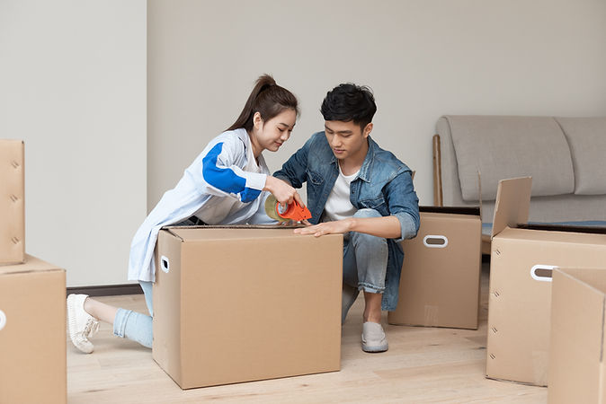 A young couple packing cardboard boxes in their living room, preparing for a move. The woman is sealing a box with tape while the man assists.