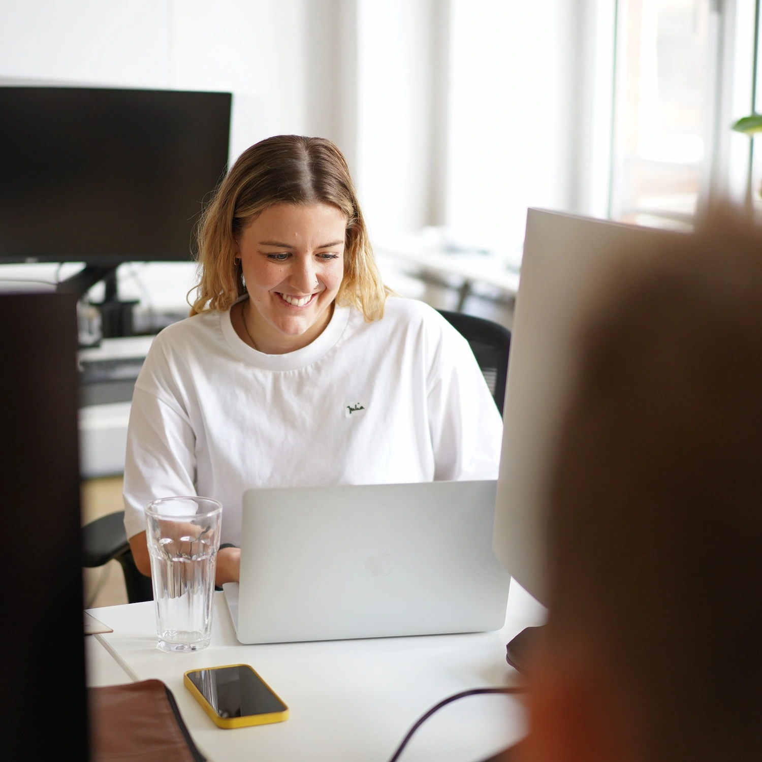Lächelnde Frau arbeitet an einem Laptop in einem hellen Büro mit Wasser-Glas und Smartphone auf dem Tisch.