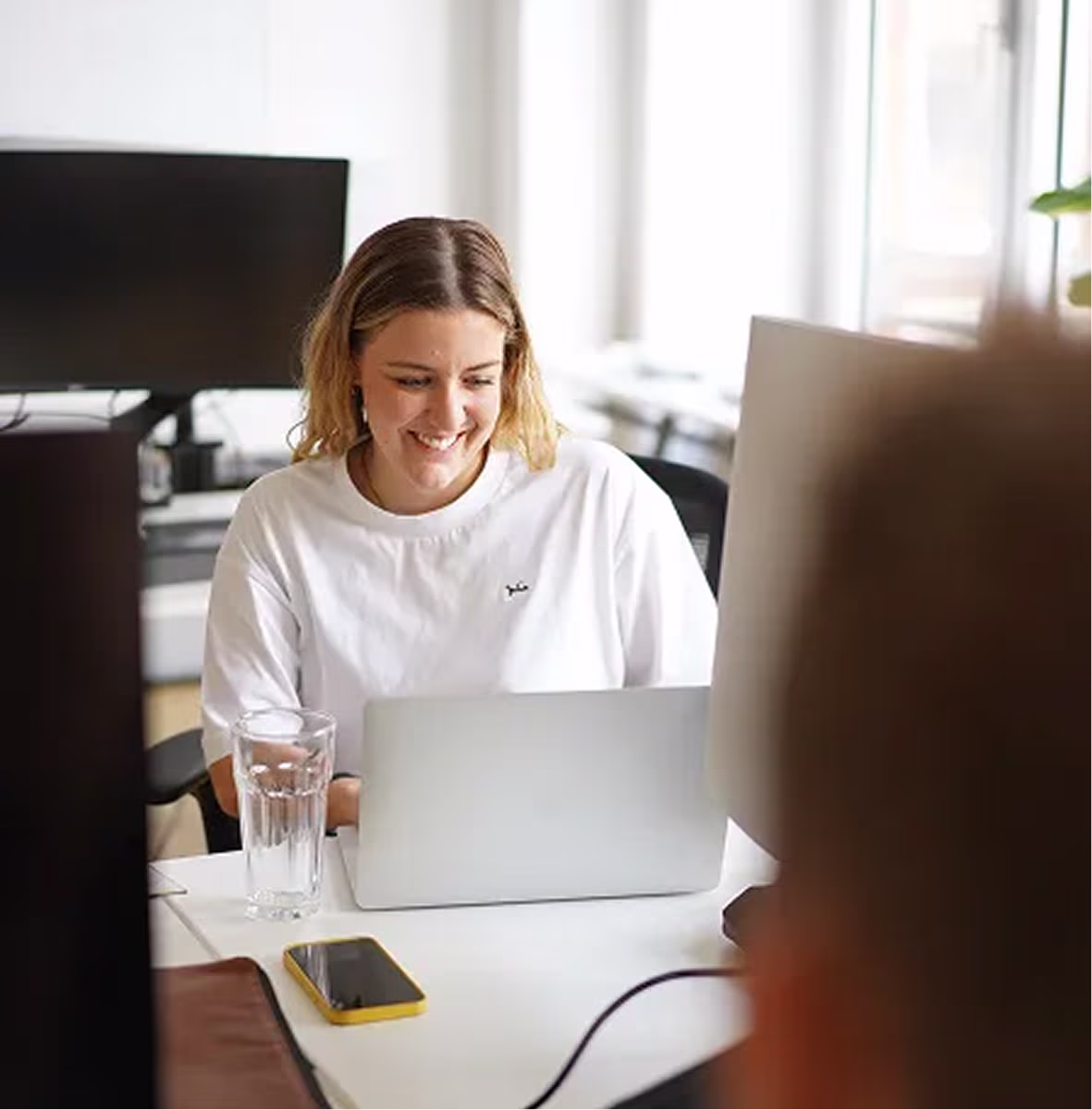 Lächelnde Frau arbeitet an einem Laptop in einem hellen Büro mit Wasser-Glas und Smartphone auf dem Tisch.