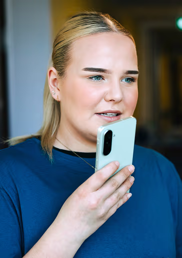 Junge Frau hält ein Smartphone nah an ihrem Mund, spricht und blickt leicht nach rechts.