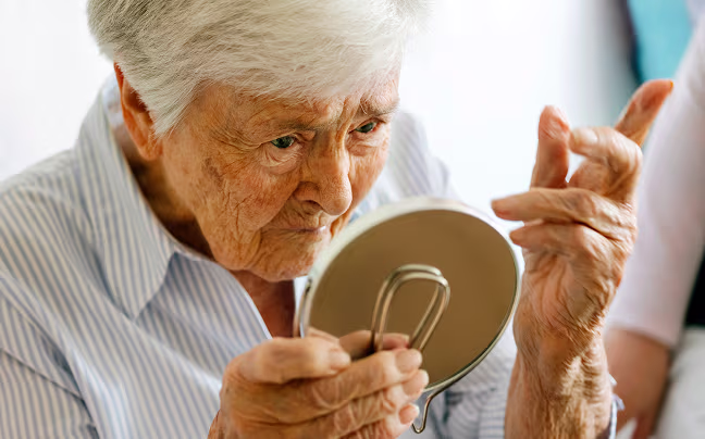 Ältere Frau mit grauen Haaren betrachtet aufmerksam ihr Gesicht in einem kleinen Handspiegel.
