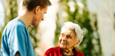 Ältere Frau mit grauen Haaren lächelt einen Mann in medizinischer Schutzkleidung an.