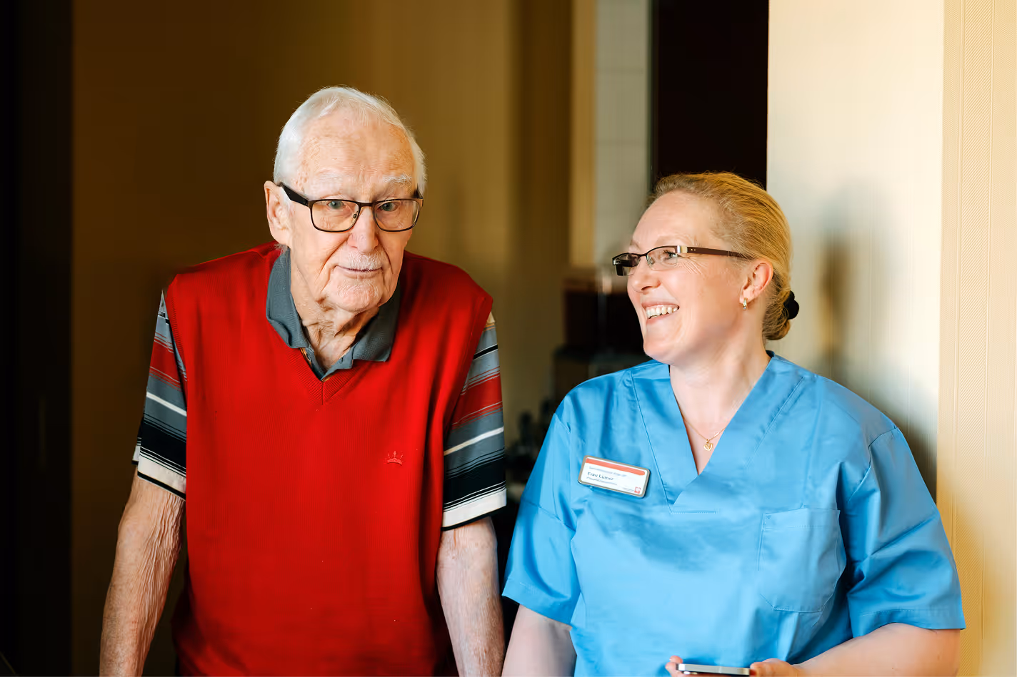 Älterer Herr mit Brille und rotem Pullover steht neben einer lächelnden Pflegekraft in blauer Uniform.