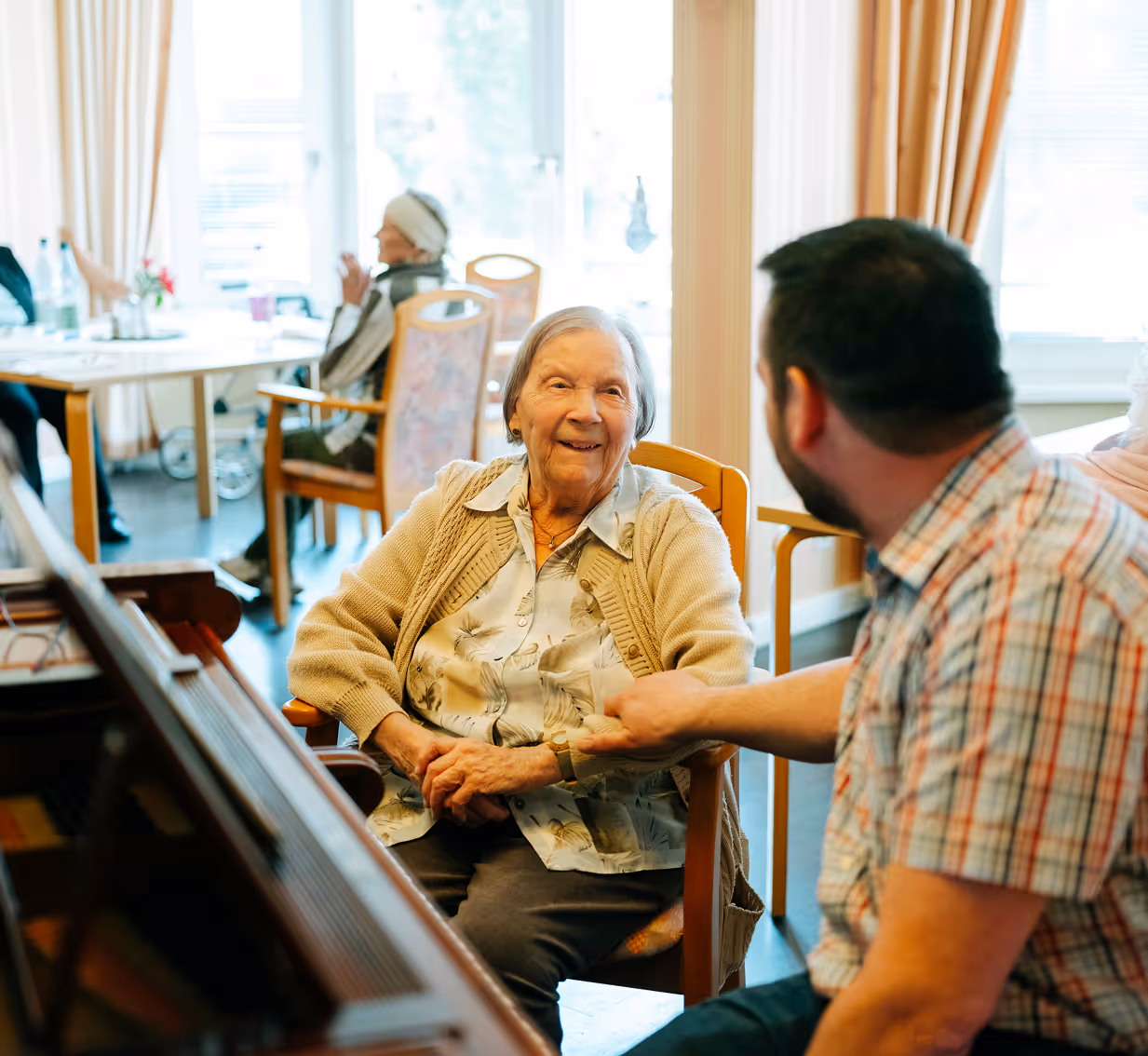 Ältere Frau sitzt am Klavier und lächelt einen Mann an, der sich zu ihr dreht.