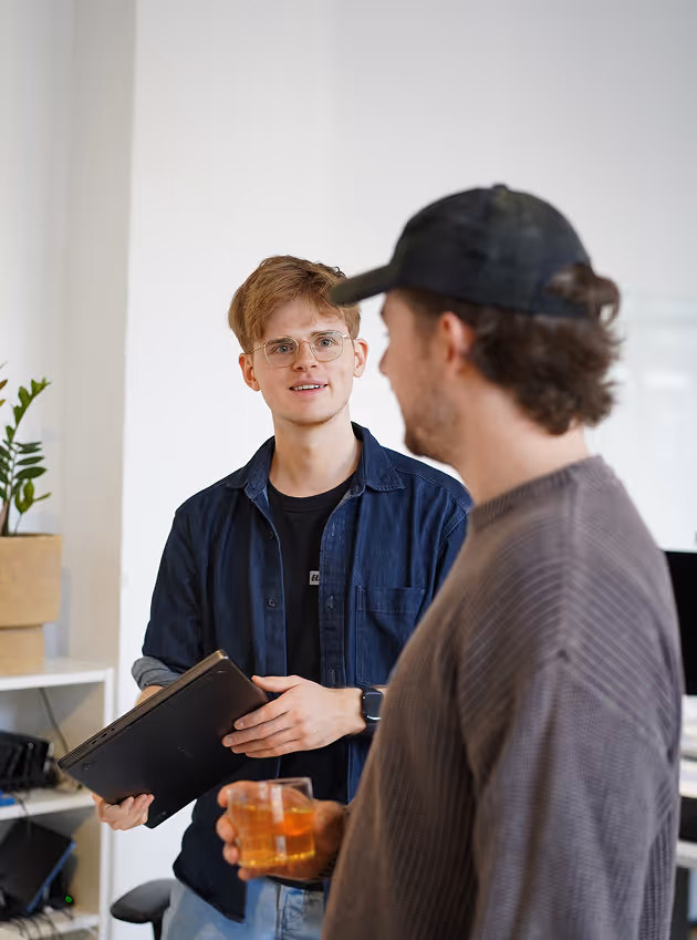 Zwei Männer unterhalten sich im Büro, einer hält einen Laptop, der andere ein Glas mit Getränk.