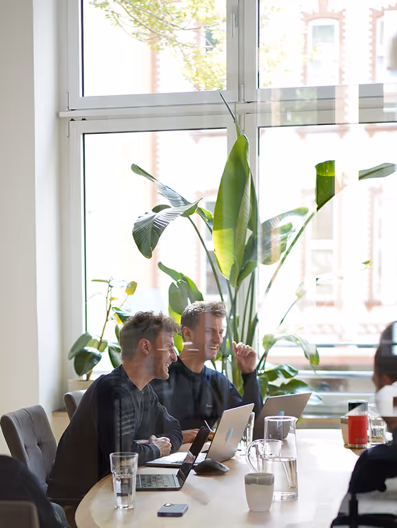 Zwei junge Männer sitzen an einem Tisch mit Laptops und Gläsern, unterhalten sich in einem hellen Raum mit großen Fenstern und grünen Pflanzen.