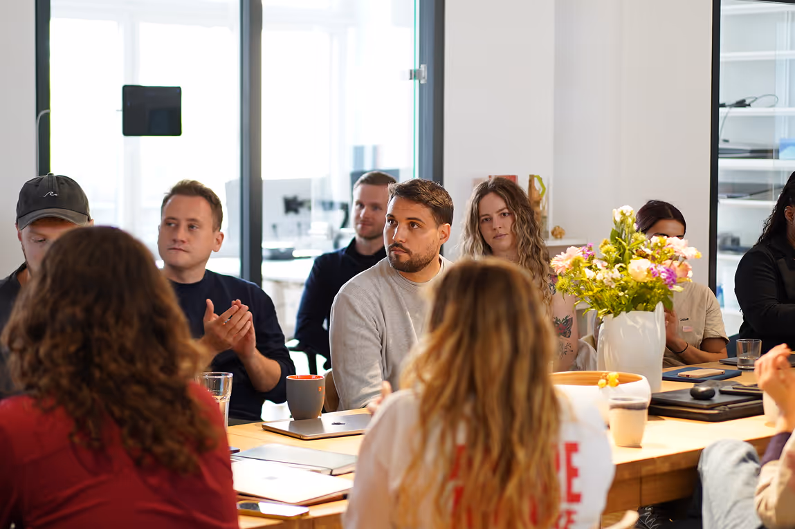 Personen in einem Besprechungsraum sitzen um einen Tisch und hören aufmerksam zu, einige klatschen.